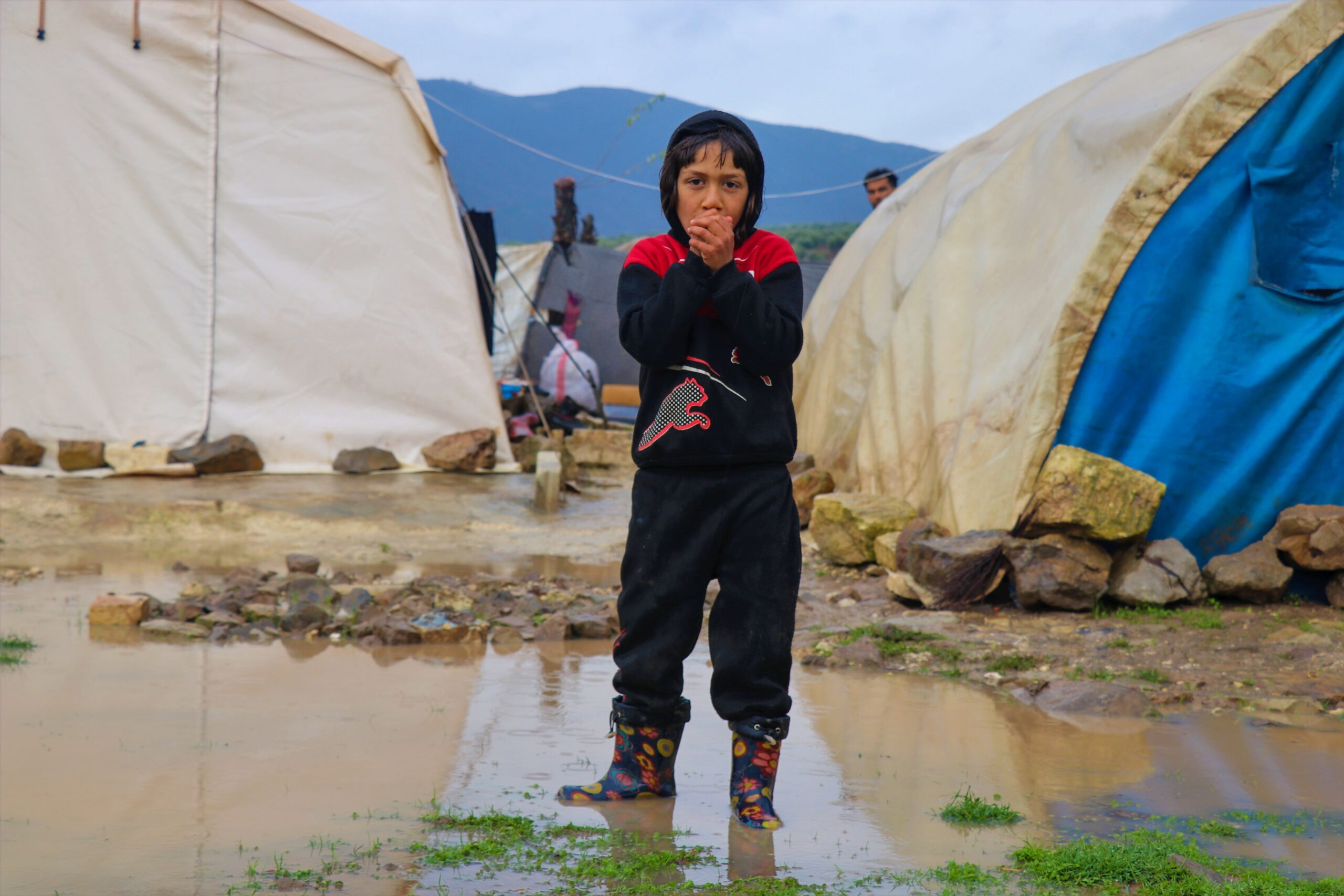 A child stands in a muddy refugee camp in Syria, appearing cold and in need of warmth.